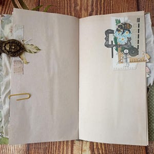 May include: Open journal with blank pages, adorned with decorative elements. A brass-toned metal clasp with a face design secures the cover. A paperclip holds pages together. Decorative paper and fabric accents add texture and visual interest. A collage with a bee and flowers is on the right page.