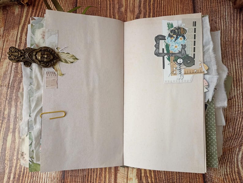 May include: Open journal with blank pages, adorned with decorative elements. A brass-toned metal clasp with a face design secures the cover. A paperclip holds pages together. Decorative paper and fabric accents add texture and visual interest. A collage with a bee and flowers is on the right page.