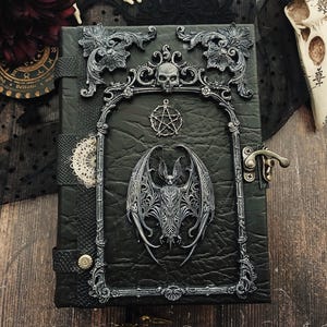 May include: A black leather-bound journal with ornate silver-colored embellishments. The cover features a bat, skull, and pentagram design. The journal has a clasp closure and decorative trim, evoking a gothic or fantasy theme.