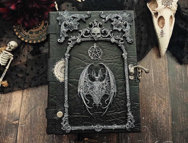 May include: A black leather-bound journal with ornate silver-colored embellishments. The cover features a bat, skull, and pentagram design. The journal has a clasp closure and decorative trim, evoking a gothic or fantasy theme.