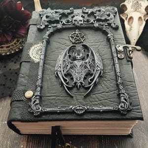 May include: A black leather-bound journal with ornate silver-colored embellishments. The cover features a bat, skull, and pentagram design. The journal has a clasp closure and a decorative frame around the bat.