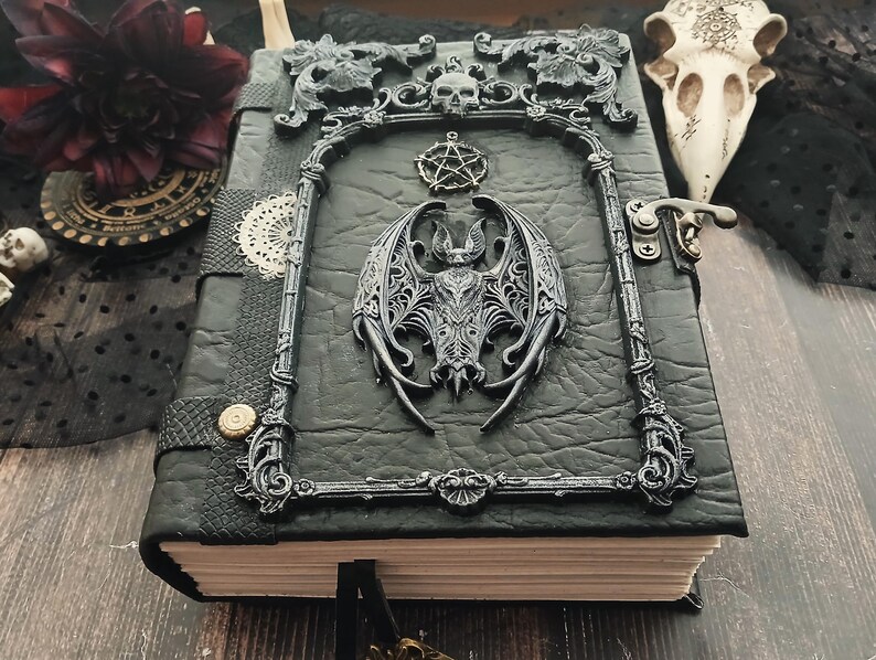 May include: A black leather-bound journal with ornate silver-colored embellishments. The cover features a bat, skull, and pentagram design. The journal has a clasp closure and a decorative frame around the bat.