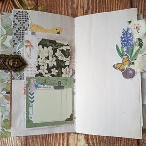 May include: Open journal with decorative pages. Features include a brass-toned metal clasp, floral and botanical paper accents, and a sticker of a butterfly and flowers. The journal is open on a wooden surface.