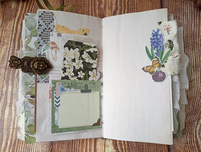 May include: Open journal with decorative pages. Features include a brass-toned metal clasp, floral and botanical paper accents, and a sticker of a butterfly and flowers. The journal is open on a wooden surface.