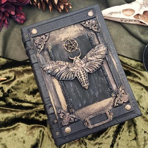 Puede incluir: Un libro oscuro y ornamentado con un dise&ntilde;o de polilla y pentagrama. La cubierta es negra con detalles dorados y esquinas decorativas. La polilla es el punto focal, con alas y cuerpo detallados. El libro tiene un tema g&oacute;tico o de fantas&iacute;a.