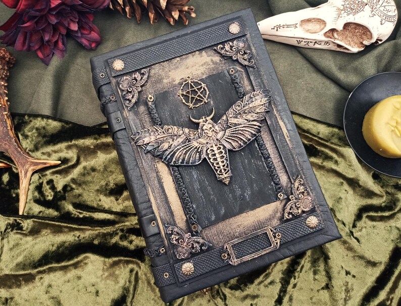 Puede incluir: Un libro oscuro y ornamentado con un dise&ntilde;o de polilla y pentagrama. La cubierta es negra con detalles dorados y esquinas decorativas. La polilla es el punto focal, con alas y cuerpo detallados. El libro tiene un tema g&oacute;tico o de fantas&iacute;a.