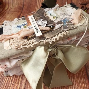 May include: A handmade journal with a soft, gray cover and a large, sage green satin bow. The journal is adorned with lace, fabric scraps, and decorative elements, including a butterfly and the text "catch a moment". The journal is on a wooden surface.