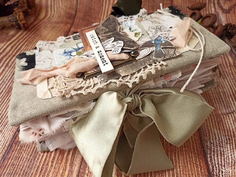 May include: A handmade journal with a soft, gray cover and a large, sage green satin bow. The journal is adorned with lace, fabric scraps, and decorative elements, including a butterfly and the text "catch a moment". The journal is on a wooden surface.