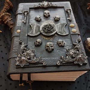 Peut inclure: Un journal gothique gris fonc&eacute; avec des ornements m&eacute;talliques &eacute;labor&eacute;s. La couverture pr&eacute;sente des motifs de cr&acirc;nes, un pentagramme et des breloques en forme de cl&eacute;s. Le journal a un aspect textur&eacute; semblable au cuir et un cadre m&eacute;tallique d&eacute;coratif.
