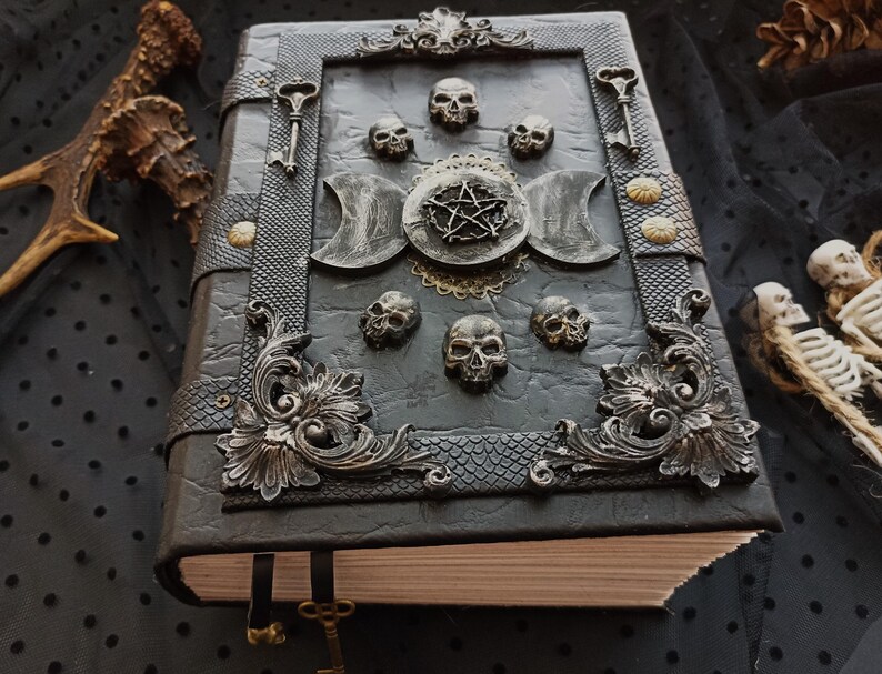 Peut inclure: Un journal gothique gris fonc&eacute; avec des ornements m&eacute;talliques &eacute;labor&eacute;s. La couverture pr&eacute;sente des motifs de cr&acirc;nes, un pentagramme et des breloques en forme de cl&eacute;s. Le journal a un aspect textur&eacute; semblable au cuir et un cadre m&eacute;tallique d&eacute;coratif.