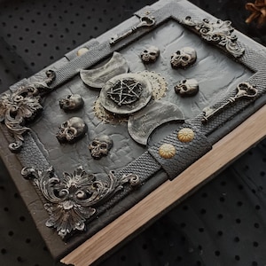 Peut inclure: Un livre d&eacute;cor&eacute; de couleur gris fonc&eacute;, orn&eacute; de cr&acirc;nes, de croissants de lune et d'un pentagramme. La couverture textur&eacute;e est encadr&eacute;e et comporte une sangle en cuir avec des clous dor&eacute;s. Le th&egrave;me g&eacute;n&eacute;ral est gothique et mystique.