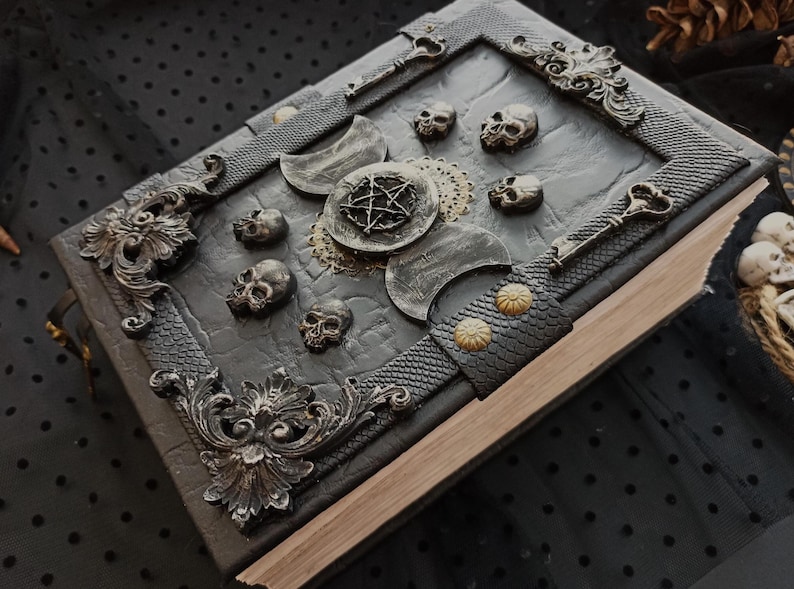 Peut inclure: Un livre d&eacute;cor&eacute; de couleur gris fonc&eacute;, orn&eacute; de cr&acirc;nes, de croissants de lune et d'un pentagramme. La couverture textur&eacute;e est encadr&eacute;e et comporte une sangle en cuir avec des clous dor&eacute;s. Le th&egrave;me g&eacute;n&eacute;ral est gothique et mystique.