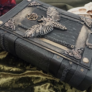 Puede incluir: Un libro oscuro de estilo g&oacute;tico con detalles intrincados. La cubierta negra presenta adornos met&aacute;licos, incluyendo una polilla y un pentagrama. Los bordes texturizados sugieren un tema m&iacute;stico o de fantas&iacute;a, en cm.