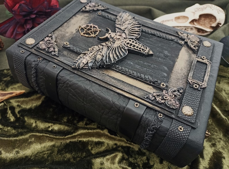 Puede incluir: Un libro oscuro de estilo g&oacute;tico con detalles intrincados. La cubierta negra presenta adornos met&aacute;licos, incluyendo una polilla y un pentagrama. Los bordes texturizados sugieren un tema m&iacute;stico o de fantas&iacute;a, en cm.