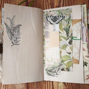 May include: Open journal with botanical illustrations. One page features a black and white drawing of lily of the valley. The facing page has a butterfly and frog sticker, with a collage of green leaves and paper. The journal is bound with a decorative clasp.