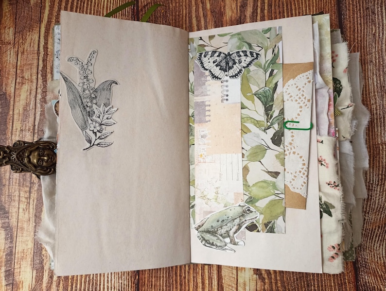 May include: Open journal with botanical illustrations. One page features a black and white drawing of lily of the valley. The facing page has a butterfly and frog sticker, with a collage of green leaves and paper. The journal is bound with a decorative clasp.