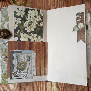 May include: Open journal with decorative pages. Features include a floral print, wood grain, and a collage with a bird's nest illustration. The journal has a bronze-colored clasp with a face design.