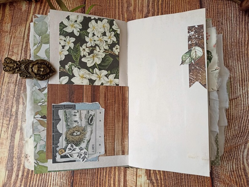 May include: Open journal with decorative pages. Features include a floral print, wood grain, and a collage with a bird's nest illustration. The journal has a bronze-colored clasp with a face design.