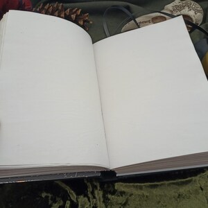 Puede incluir: Un diario abierto con p&aacute;ginas en blanco de color crema. El libro est&aacute; encuadernado en negro y tiene un cierre decorativo. El diario se muestra sobre una superficie de terciopelo verde oscuro, con otros elementos decorativos en el fondo.