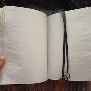May include: Open journal with blank, textured pages. The cover has a black and gold pattern. A black ribbon with a pentacle charm and a small orange flower serves as a bookmark. The journal is open, revealing the pages.