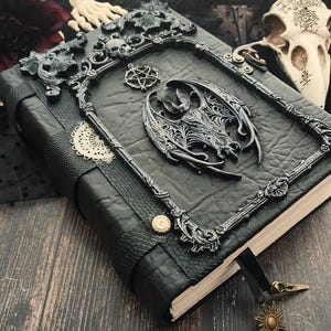 May include: A black leather-bound journal with ornate metal accents. The cover features a decorative frame with skulls and a central dragon motif. A pentagram charm and sun-shaped bookmark add detail to this gothic-themed notebook.