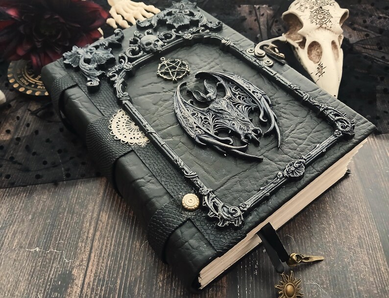 May include: A black leather-bound journal with ornate metal accents. The cover features a decorative frame with skulls and a central dragon motif. A pentagram charm and sun-shaped bookmark add detail to this gothic-themed notebook.