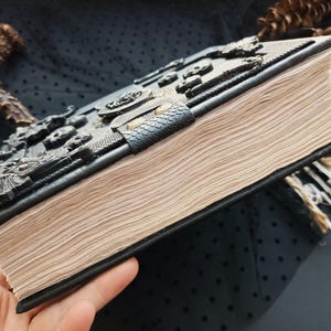 Peut inclure: Un livre reli&eacute; en cuir noir avec un motif en relief. Le livre a une couverture textur&eacute;e avec des d&eacute;tails en relief complexes et un fermoir. Les pages sont vieillies, et un marque-page de petits squelettes est visible.