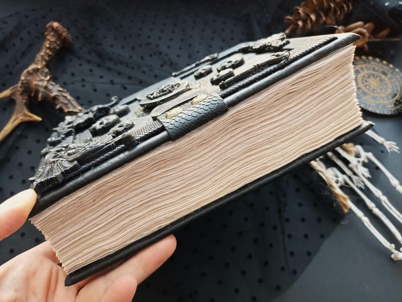 Peut inclure: Un livre reli&eacute; en cuir noir avec un motif en relief. Le livre a une couverture textur&eacute;e avec des d&eacute;tails en relief complexes et un fermoir. Les pages sont vieillies, et un marque-page de petits squelettes est visible.