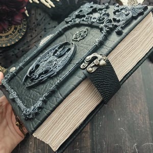 May include: A black, gothic-style journal with ornate, raised embellishments. The cover features a sculpted dragon and decorative frame. A black leather strap with a metal clasp secures the aged, cream-colored pages. The journal has a dark, textured appearance.