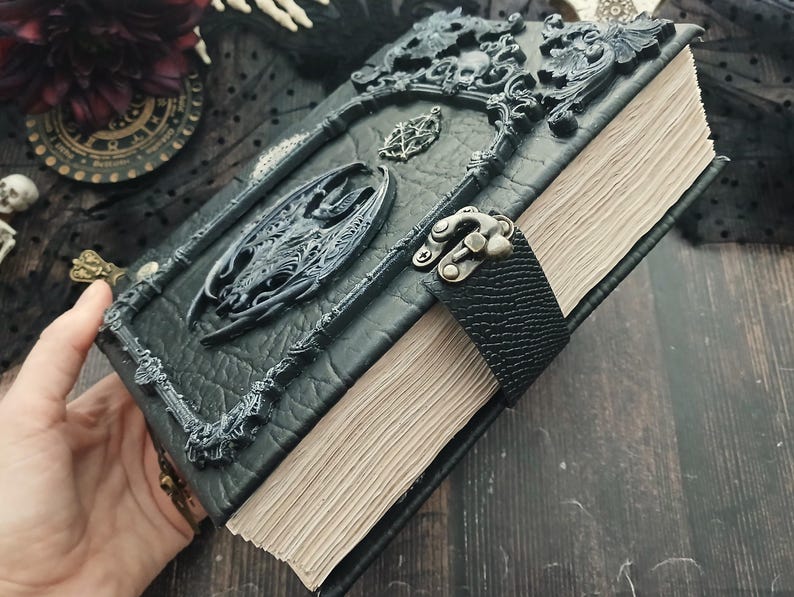 May include: A black, gothic-style journal with ornate, raised embellishments. The cover features a sculpted dragon and decorative frame. A black leather strap with a metal clasp secures the aged, cream-colored pages. The journal has a dark, textured appearance.