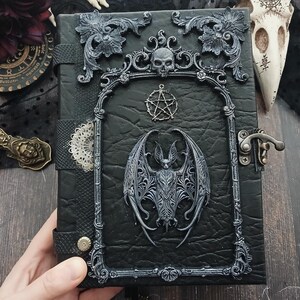 May include: Black leather-bound journal with ornate silver-colored embellishments. Features a bat, skull, and pentagram design. Includes a metal clasp closure and decorative frame. Gothic-themed notebook.