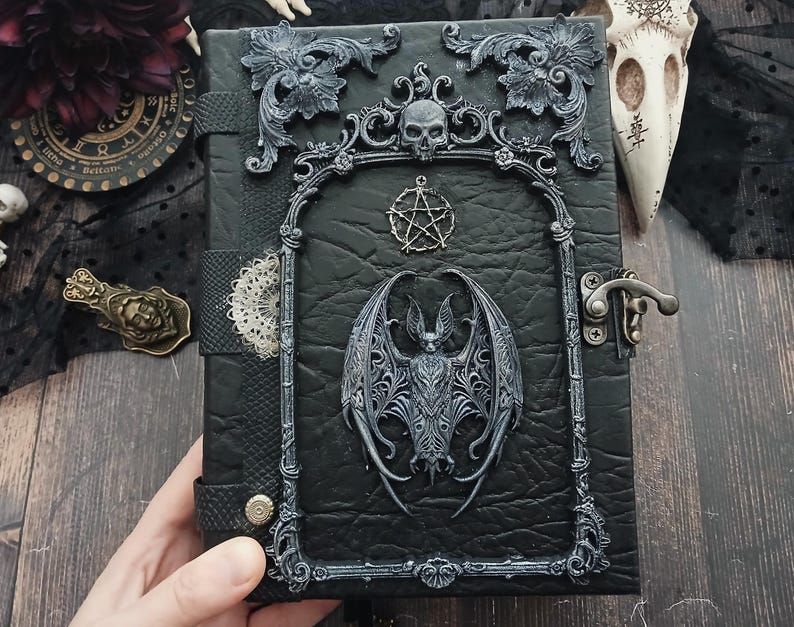 May include: Black leather-bound journal with ornate silver-colored embellishments. Features a bat, skull, and pentagram design. Includes a metal clasp closure and decorative frame. Gothic-themed notebook.