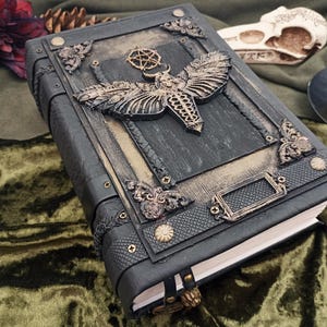 Puede incluir: Un diario de cuero gris oscuro con adornos met&aacute;licos ornamentados. La cubierta presenta un gran dise&ntilde;o de polilla y piezas decorativas en las esquinas. El diario tiene un cierre de lat&oacute;n y una cinta de marcador. El tema general es g&oacute;tico y fantas&iacute;a.