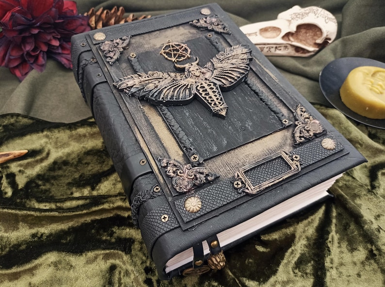 Puede incluir: Un diario de cuero gris oscuro con adornos met&aacute;licos ornamentados. La cubierta presenta un gran dise&ntilde;o de polilla y piezas decorativas en las esquinas. El diario tiene un cierre de lat&oacute;n y una cinta de marcador. El tema general es g&oacute;tico y fantas&iacute;a.