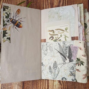 May include: Open journal with decorative pages. A large bee sticker is on the left page. The right page has a pocket with a botanical illustration and a ticket. The journal is bound with a decorative clasp and fabric edging.