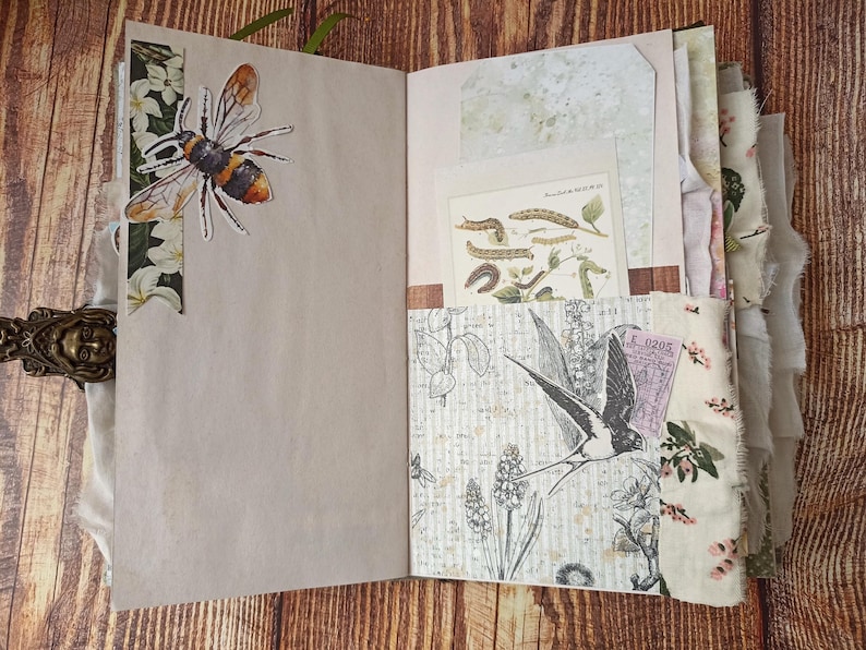 May include: Open journal with decorative pages. A large bee sticker is on the left page. The right page has a pocket with a botanical illustration and a ticket. The journal is bound with a decorative clasp and fabric edging.