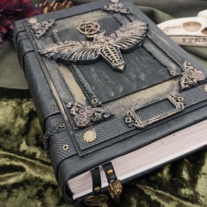 Puede incluir: Un diario de cuero gris oscuro con adornos met&aacute;licos ornamentados. La portada presenta un dise&ntilde;o detallado de polilla y piezas decorativas en las esquinas. Un marcap&aacute;ginas de cuero con un dije dorado cuelga de las p&aacute;ginas. El diario descansa sobre terciopelo verde.