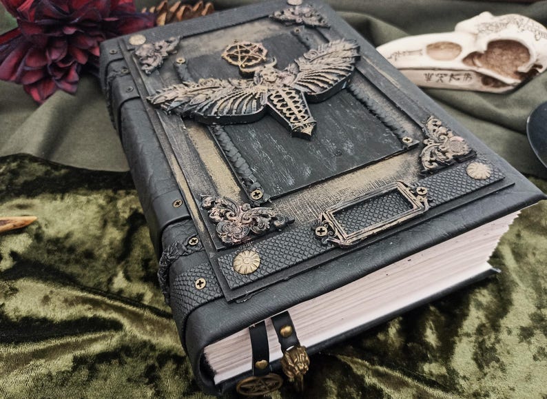 Puede incluir: Un diario de cuero gris oscuro con adornos met&aacute;licos ornamentados. La portada presenta un dise&ntilde;o detallado de polilla y piezas decorativas en las esquinas. Un marcap&aacute;ginas de cuero con un dije dorado cuelga de las p&aacute;ginas. El diario descansa sobre terciopelo verde.