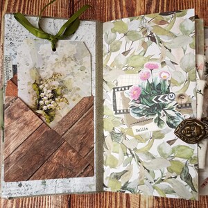 May include: Open journal with decorative pages featuring floral and wood-grain patterns. A tag with a bouquet of white flowers is tucked into a wooden pocket. The journal is bound with a green ribbon and a decorative clasp.