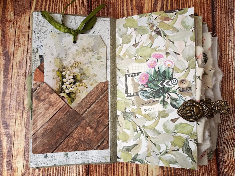 May include: Open journal with decorative pages featuring floral and wood-grain patterns. A tag with a bouquet of white flowers is tucked into a wooden pocket. The journal is bound with a green ribbon and a decorative clasp.