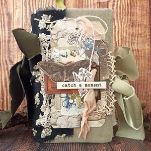 May include: A handmade journal with a fabric cover in shades of green and black, adorned with lace, tags, and a ribbon. The journal features a butterfly illustration and the text "catch a moment".