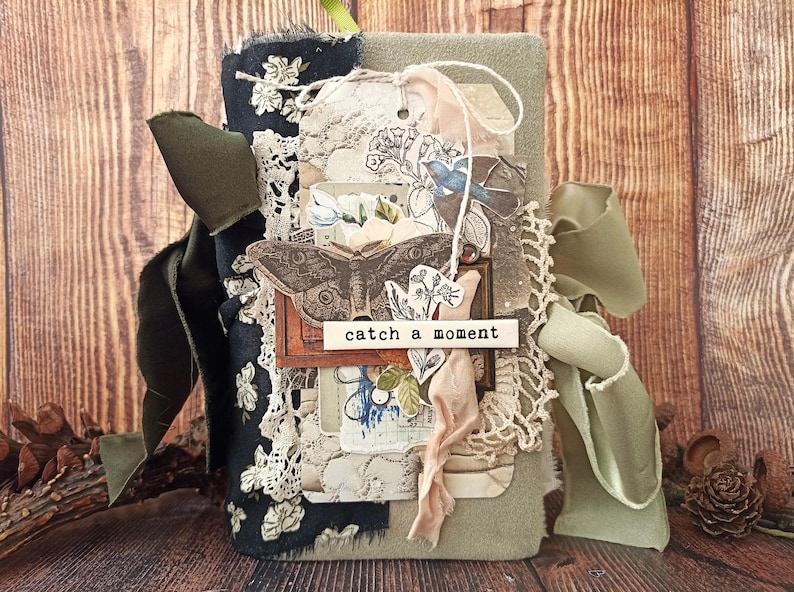 May include: A handmade journal with a fabric cover in shades of green and black, adorned with lace, tags, and a ribbon. The journal features a butterfly illustration and the text "catch a moment".