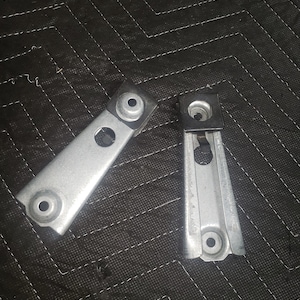 May include: Two silver metal brackets with holes and a black rubber piece on one of the brackets.