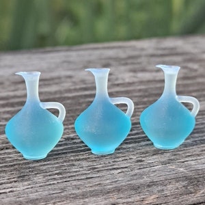 May include: Three small, light blue glass jugs with handles. The jugs are frosted and have a slightly iridescent finish.