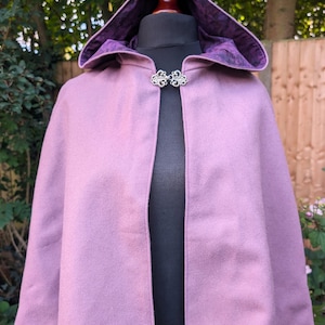 May include: A short, mauve-colored hooded cape featuring a smooth, solid-colored exterior and a dark purple, patterned lining. The garment is secured at the neckline with an ornate, silver-toned metal clasp featuring intricate scrollwork details.