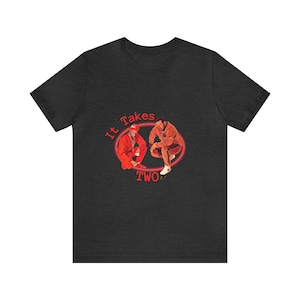 May include: Dark grey t-shirt with a graphic of two figures in red and brown outfits, within a red oval, and the text "It Takes TWO". The design is centrally placed on the shirt.