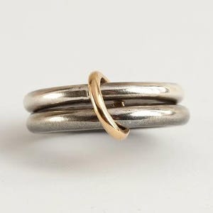 Könnte beinhalten: Ein silberfarbener Ring mit zwei Bändern, die von einem goldfarbenen Band gekreuzt werden. Die Ringe werden auf weißem Hintergrund dargestellt. Die Bänder haben eine glatte, polierte Oberfläche und erzeugen eine moderne und minimalistische Ästhetik.