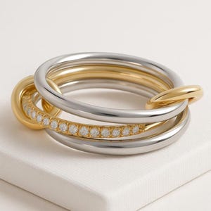 May include: A multi-tone ring set featuring gold and silver bands. One gold band is encrusted with small, clear gemstones. The rings are interlocked, creating a layered effect. The jewelry is displayed on a white surface.