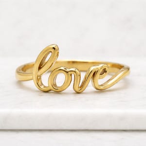 May include: A gold-colored ring with the word "love" in cursive script. The ring is made of a shiny, polished metal and sits on a white surface. The ring's design is simple yet elegant, making it a perfect accessory.