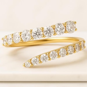 Puede incluir: Un anillo dorado envolvente con una serie de diamantes redondos y brillantes. El diseño del anillo se enrolla alrededor del dedo, con los diamantes dispuestos en una línea continua. El anillo se muestra sobre una superficie blanca.